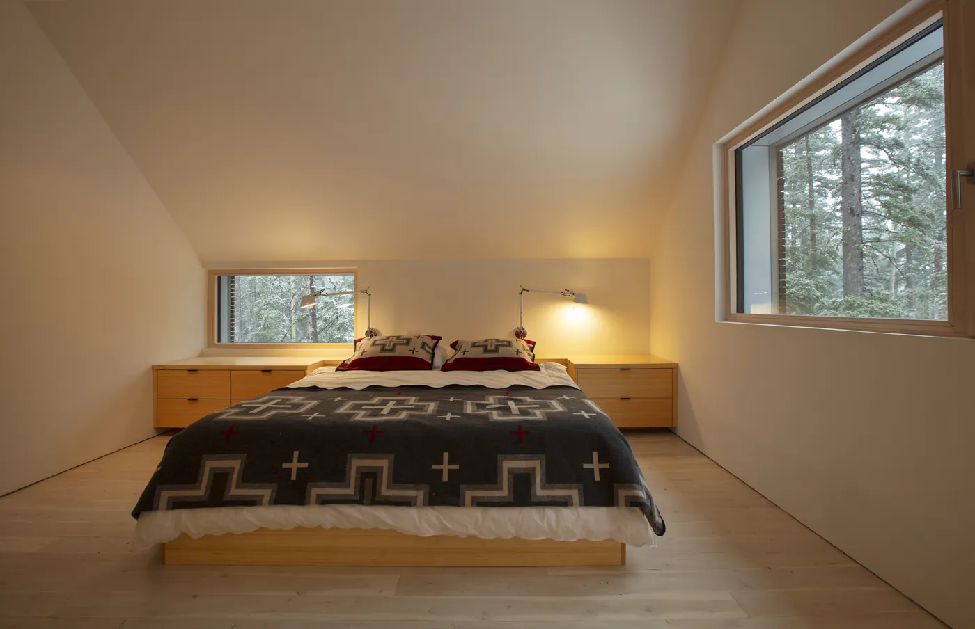 A 9k House bedroom under a raked ceiling, wood-framed windows onto snowy pines either side of the bed