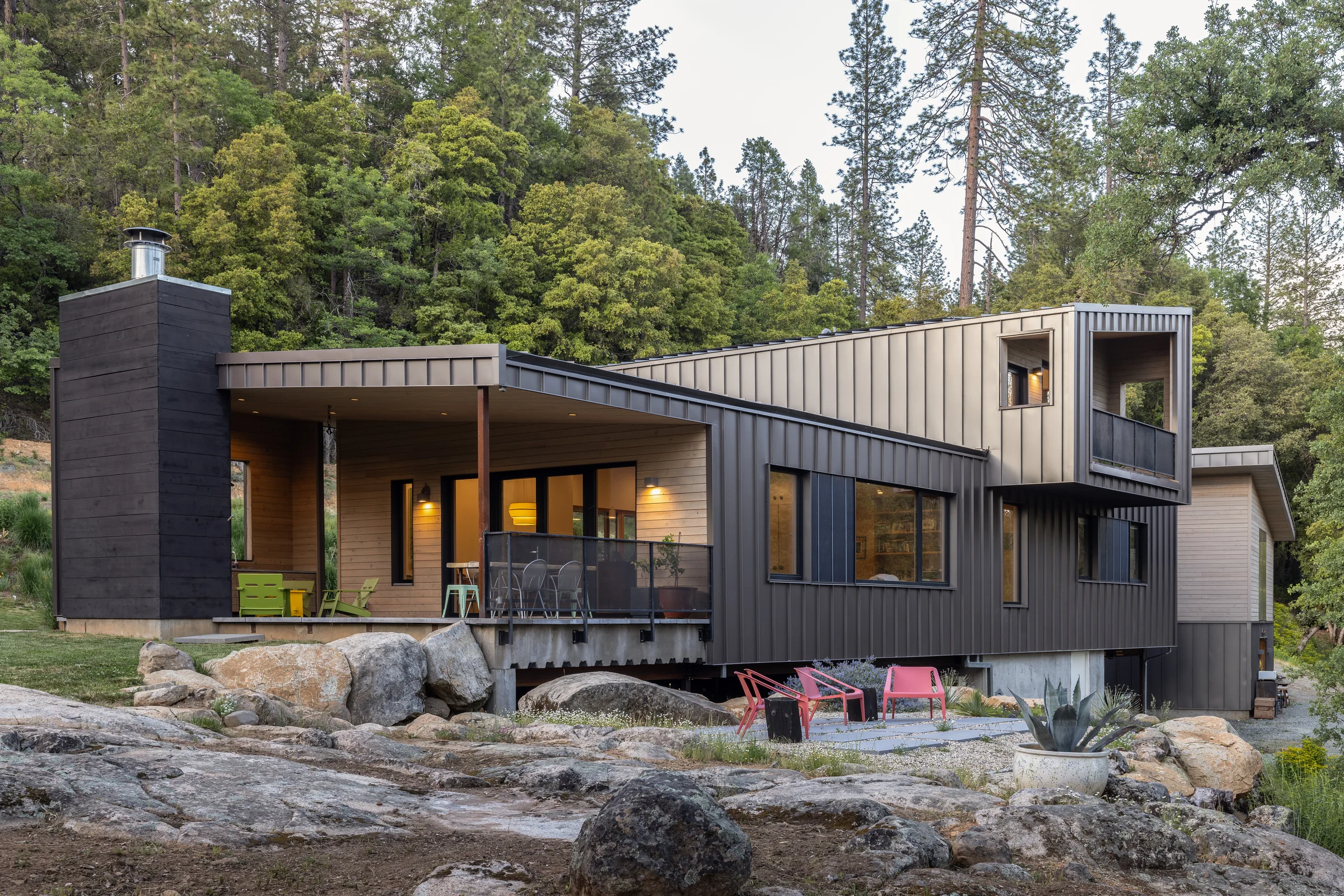 Good Haus at dusk, a dark standing-seam volume stepping down a granite slope with a lit porch and pines behind