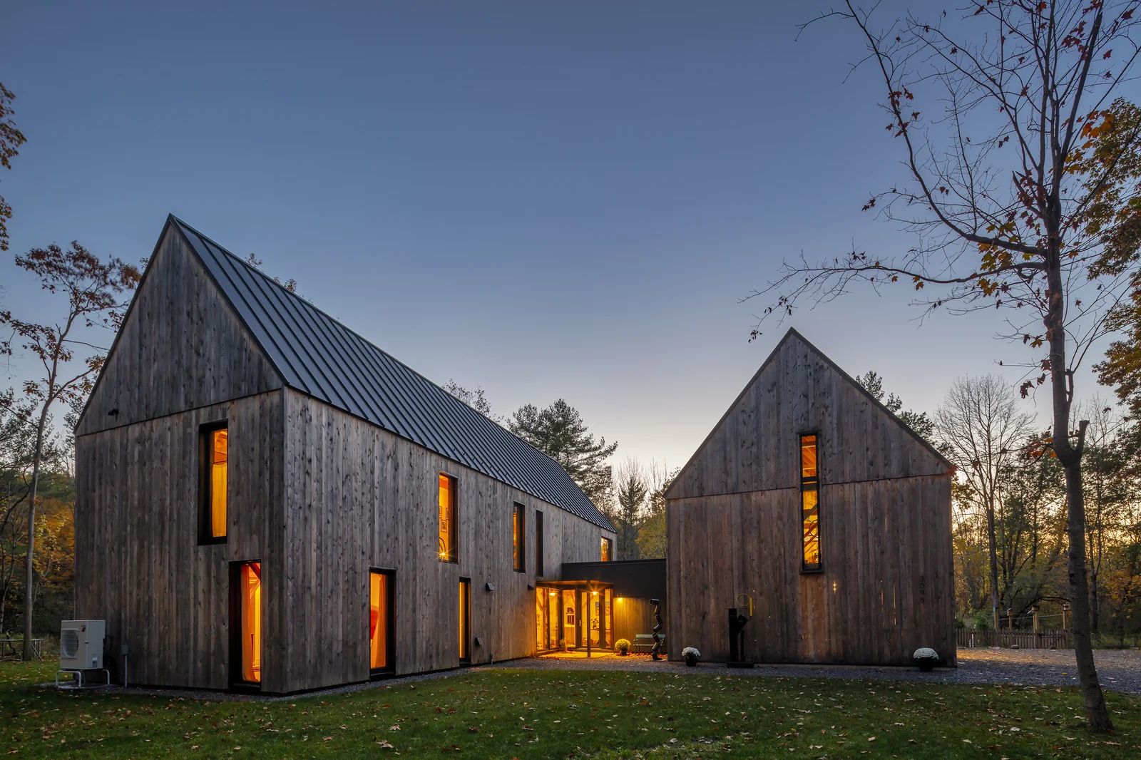 House in the Woods at dusk — a pair of dark-stained gabled volumes with Zola windows lit from within