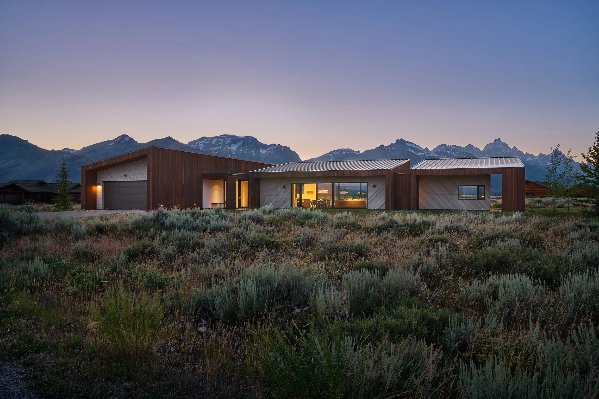 House of Fir at dusk below the Tetons, its Zola windows lit across sagebrush